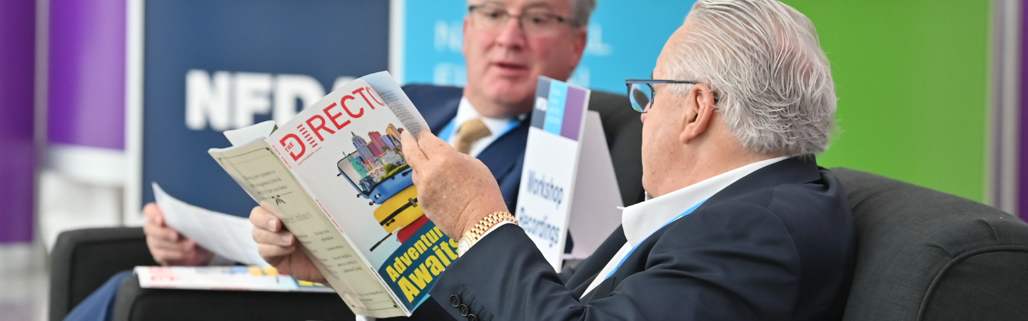 Two men in suits, seated and talking while looking at a magazine.