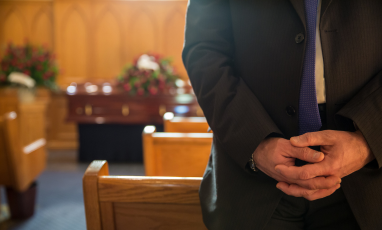 Person in a suit standing in front of a casket with flowers.